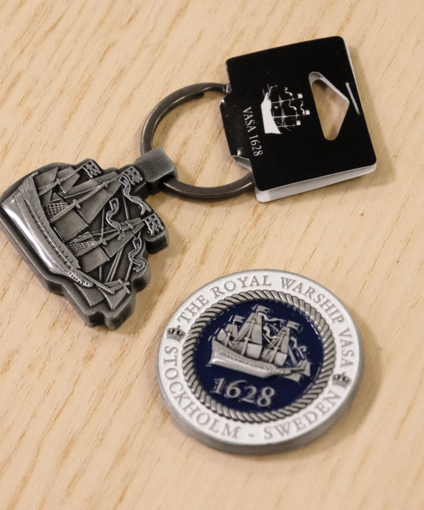 En metallf&auml;rgad Vasa &ouml;rlogsskepp-nyckelring och en rund Vasa 1628-magnet med bl&aring; emalj, utlagda p&aring; ett tr&auml;underlag